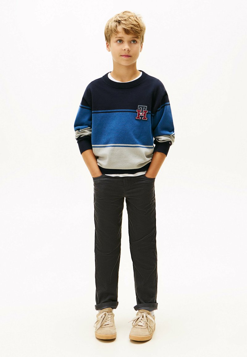 Jongen in een blauwgrijs gestreept sweater, donkere broek en beige sneakers, staand met handen in zijn zakken tegen een witte achtergrond.