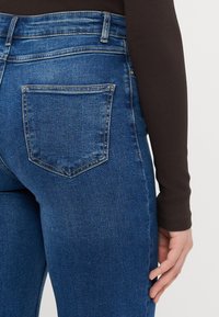 Temno modri traper hlače s prilegajočo zasnovo, ki vključujejo zadnji žep in gladko teksturo. Pas je strukturiran in visok.