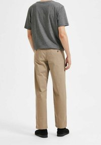 Šedé tričko s krátkým rukávem v kombinaci s béžovými kalhotami chinos. Kalhoty mají volný střih a jemné logo detail nad zadní kapsou. Jsou nošeny černé boty.