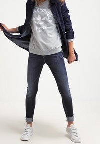 Person som bär en mörkblå jacka, grå Adidas Originals sweatshirt, mörka uppvikta tajta jeans och vita sneakers mot en neutral bakgrund.