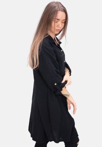 Schwarzes Hemdblusenkleid aus strukturiertem Stoff, mit hochgerollten langen Ärmeln und silbernen Knöpfen, das eine lässige Silhouette und einen knielangen Schnitt bietet.