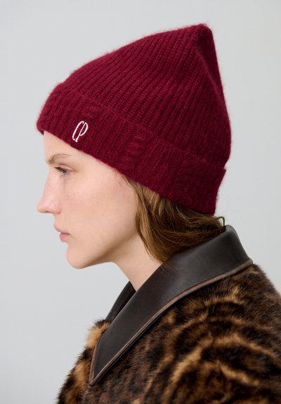 Gorro de punto burdeos con una parte superior puntiaguda, textura acanalada y un pequeño logo blanco bordado. Lleva puesto un abrigo de piel marrón con un cuello de cuero.