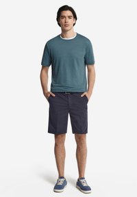 En tealfärgad bomulls t-shirt med vit rund halsringning, matchad med marinblå shorts. Modellen bär blå mockaskor med krämfärgade snörningar.