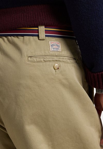 Chinos en coton beige avec une étiquette en patch, dotés d'un bouton unique et d'une poche arrière. La ceinture présente des rayures multicolores.