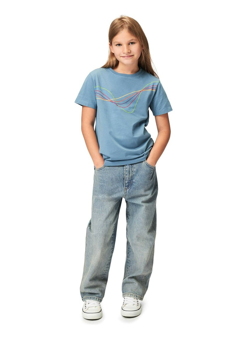 Blå bomullströja med flerfärgad vågmönster, tillsammans med stora ljusblå denimjeans och vita sneakers.