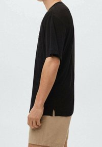 Sideprofil af person iført sort kortærmet skjorte med lomme og beige shorts mod en ensfarvet lys baggrund.