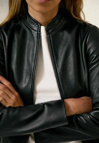 Veste aviateur en cuir noir avec un col montant, une fermeture éclair à l'avant et une texture lisse, portée par-dessus un haut blanc côtelé. Les bras sont croisés.