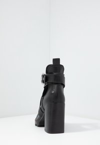 Botín de cuero negro con tacón, que presenta un tacón ancho, correa ajustable con hebilla plateada y diseño lateral con corte.