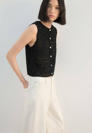 Top corto negro sin mangas con un diseño texturizado de punto y botones en la parte delantera. Combinado con pantalones blancos de pierna ancha y cintura alta.