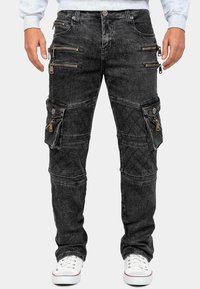 Pantaloni cargo in denim nero con diverse tasche con cerniera, texture trapuntata e fit affusolato. Gli accenti includono dettagli in metallo argentato e cuciture.