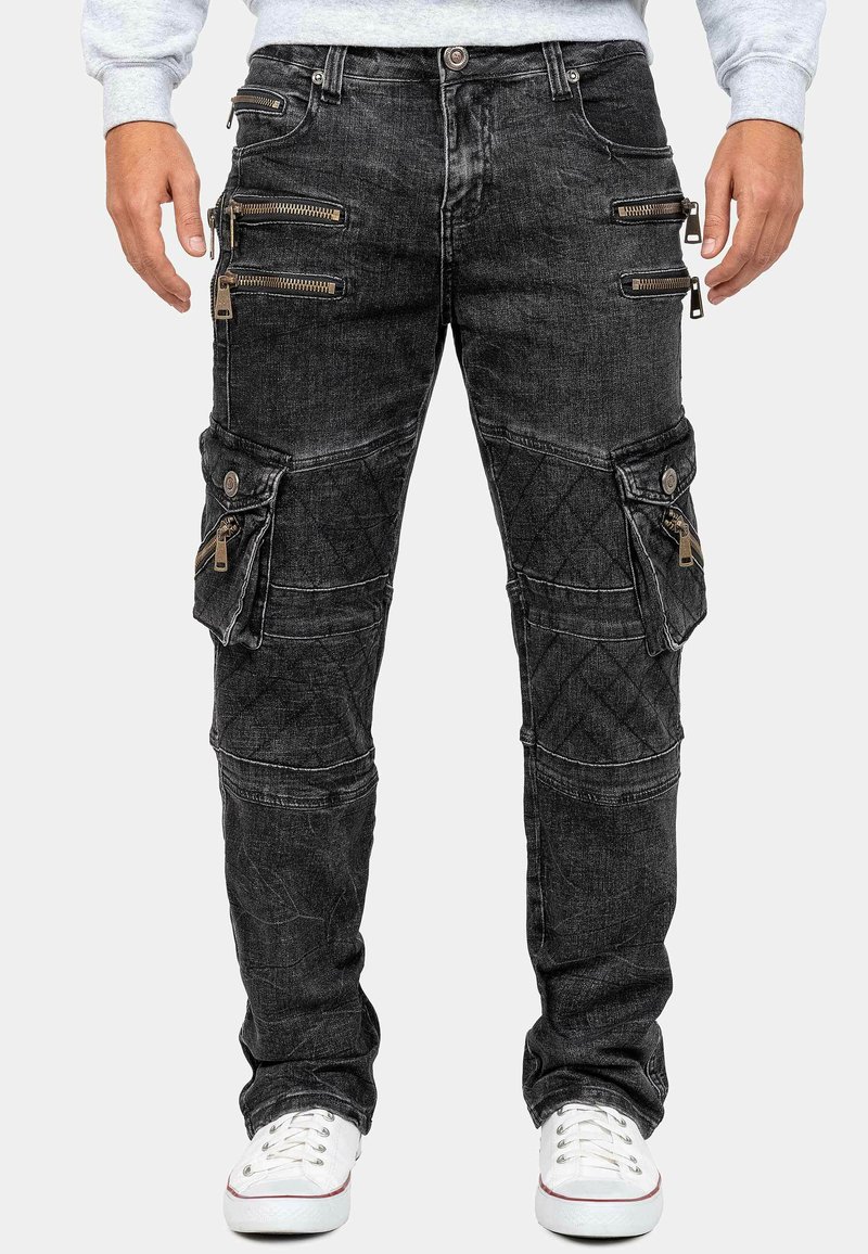 Pantaloni cargo in denim nero con diverse tasche con cerniera, texture trapuntata e fit affusolato. Gli accenti includono dettagli in metallo argentato e cuciture.