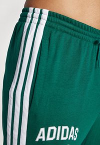 Zöld sportnadrág pamut keverékből, fehér csíkokkal az oldalán, az "ADIDAS" márkanevének fehér hímzéssel a fronton.