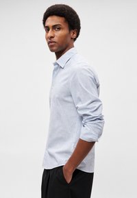 Ανοιχτό μπλε, μακρυμάνικο πουκάμισο με κουμπιά και μυτερό γιακά, μαλακή υφή και μανίκια γυρισμένα, φορεμένο με τα χέρια στις τσέπες.