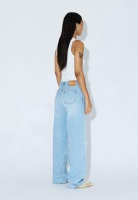 Light blue wide-leg jeans, denim fabric, high waist, five-pocket design, and brown leather tag at the back. Worn with a white tank top.