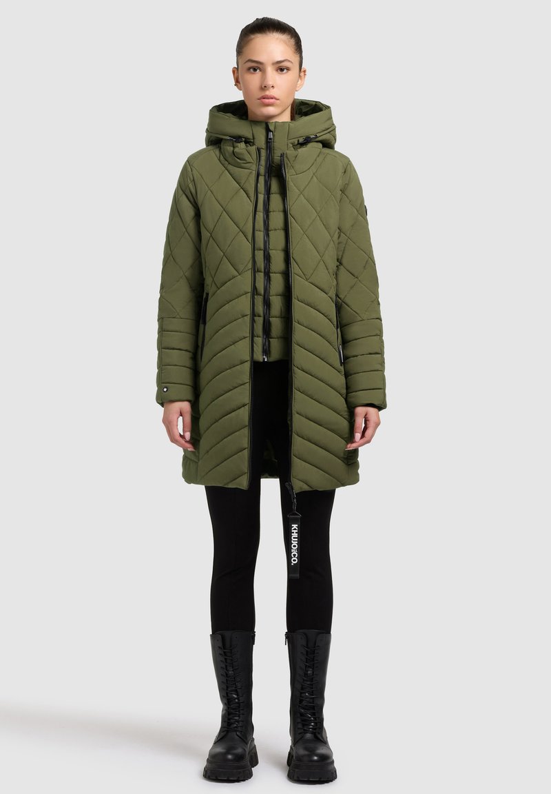 Cappotto imbottito verde oliva con cappuccio, caratterizzato da cuciture diagonali e a chevron, chiusura frontale con zip e tasche laterali. Accenti e etichette neri.