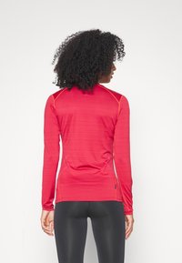 Punainen pitkähihainen urheilupaita, jossa on slim fit -muotoilu, kontrastivärisiä oransseja ompeleita, teksturoitua kangasta ja selkäsaumaa korostavaa yksityiskohtaa.