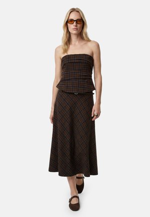 Kobieta ubrana w brązowy kratowany top bez ramiączek oraz pasującą spódnicę midi o linii A, brązowe buty typu Mary Jane i bursztynowe okulary przeciwsłoneczne, stojąca na białym tle.