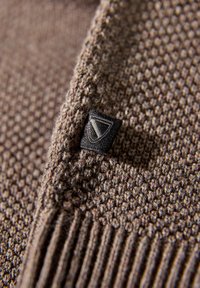 Brązowa dzianina o teksturowanej powierzchni posiada prążkowaną bordiurę oraz małą czarną naszywkę z trójkątnym wzorem z boku.
