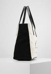 Torba na ramię w kremowym i czarnym płótnie z czarnymi skórzanymi paskami. Posiada nadruk logo oraz przestrzenny prostokątny kształt.