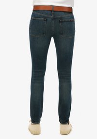 Jeans ajustados de denim azul oscuro con una textura suave, que cuentan con dos bolsillos traseros y un cinturón marrón. Terminados con costuras claras y un branding sutil.