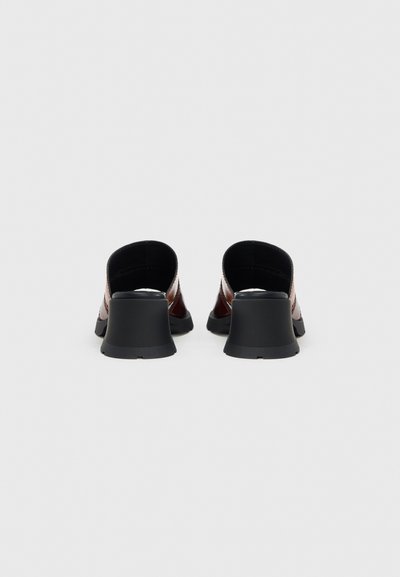 Καφέ δερμάτινα παπούτσια χωρίς κορδόνια με καμπυλωτή μαύρη φτέρνα, με λεία υφή και μινιμαλιστικό σχεδιασμό, φαίνονται από πίσω.