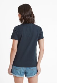Woman with shoulder-length brown hair wearing a plain dark navy t-shirt and rolled-up blue denim shorts, standing with her back to the camera.