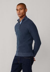 Maglione blu navy con zip e collo alla francese, design in maglia texturizzata, silhouette aderente e polsini e orlo a costine. Indossato sopra una camicia azzurra chiara e jeans scuri.