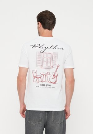 Άνδρας που φοράει λευκό T-shirt με σχέδιο παράθυρο, καρέκλα, τραπέζι, κιθάρα και το κείμενο "Rhythm" και "Good music where sound meets soul" στην πλάτη.
