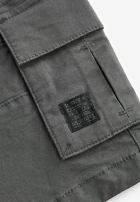 Tissu gris avec un patch noir ; l'étiquette présente des coordonnées et des informations sur l'édition ; la texture semble lisse et durable.
