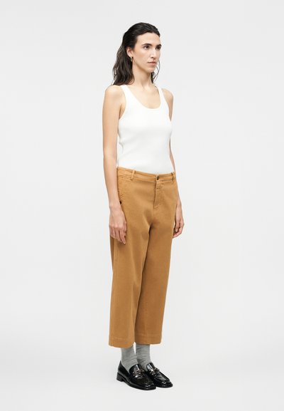 Μια γυναίκα στέκεται φορώντας λευκό αμάνικο τοπ, καφέ παντελόνι cropped, γκρι κάλτσες και μαύρα μοκασίνια, μπροστά σε απλό φωτεινό φόντο.