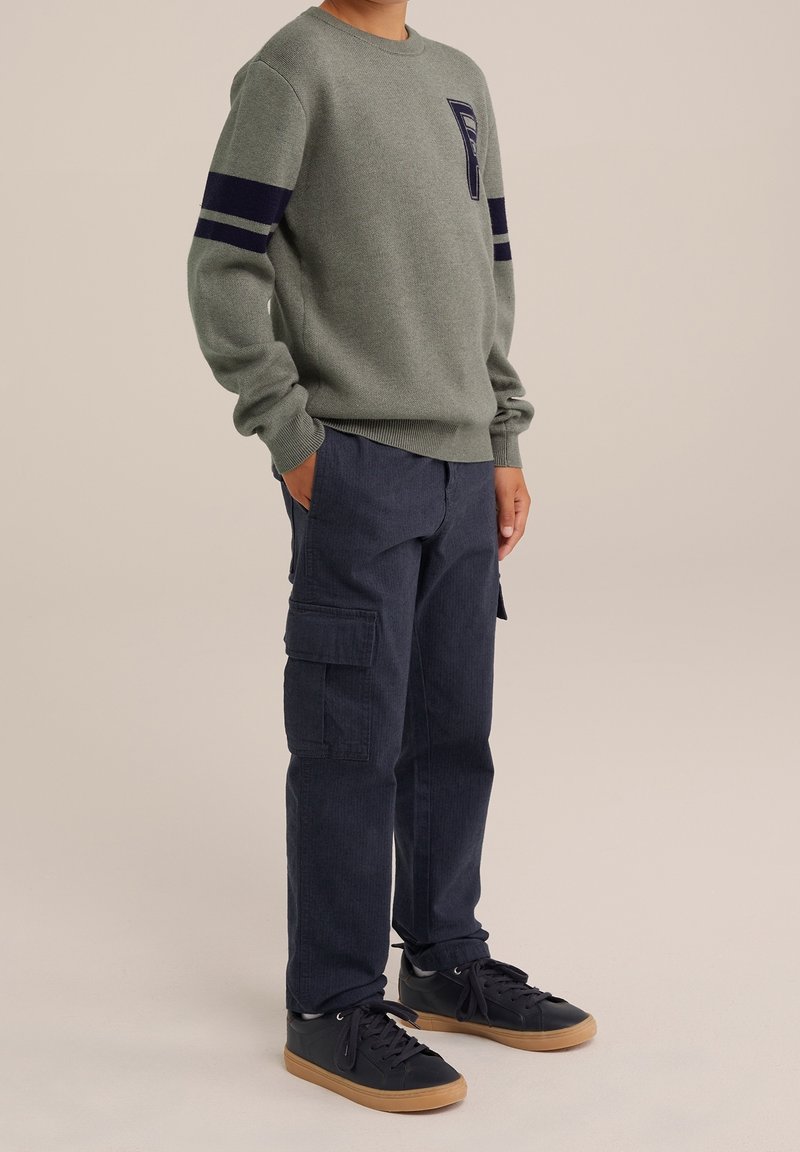Personne portant un pull gris avec des rayures bleu marine sur les manches, un pantalon cargo foncé et des baskets noires à semelles gomme, debout les mains dans les poches.