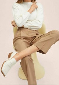 Pull à manches longues côtelé blanc, pantalon plissé beige taille haute et bottines blanches à talon carré. Fond de chaise de couleur crème.