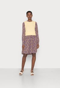 Suéter de malha creme sem mangas por cima de um vestido floral roxo de mangas longas. O vestido tem uma bainha rodada e apresenta pequenas flores amarelas e brancas.
