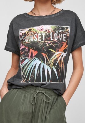 Person iført en mørkegrå t-shirt med et farverigt tropisk planteprint og teksten "SUNSET LOVE", kombineret med olivengrønne bukser med bindebånd.