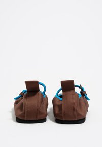 Par de zapatos de tela marrones tipo slip-on con ribetes azules y cordones ajustables azules, vistos desde la parte de atrás sobre un fondo blanco.