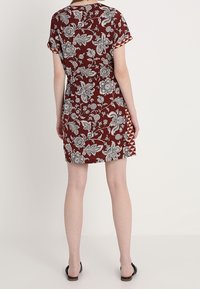 Robe bordeaux avec un motif floral blanc et gris, manches courtes et une coupe cintrée. Présente une bordure géométrique unique le long de l'ourlet.