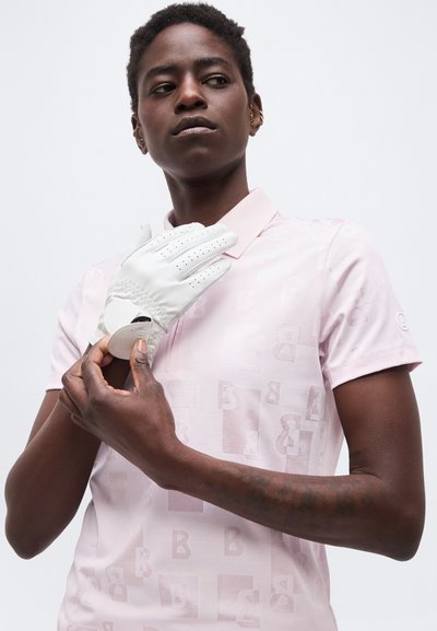 Personne portant un polo rose clair à motifs enfilant un gant blanc perforé sur la main gauche.