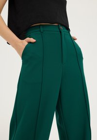 Personne portant un pantalon vert foncé taille haute à jambes larges, les mains dans les poches, et un haut noir court sur un fond clair.
