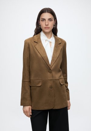 Femme aux cheveux foncés portant un blazer en daim marron sur un chemisier blanc à col noué, debout devant un fond clair uni.