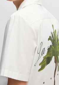 Wit shirt met korte mouwen van katoen, met een groene palmboomafbeelding en zwarte cursieve tekst op de rug. Gladde textuur.