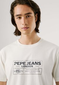 Krēmkrāsas T-krekls ar melnu grafiku, kas iekļauj tekstu "PEPE JEANS LONDON" un apļa logo, ar klasisku apaļo kakla dizainu.