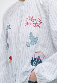 Veste blanche à rayures avec texte rouge brodé "I Run My Own Show" et images d’un hippocampe, d’un oiseau, d’un champignon et d’une théière, main dans la poche.