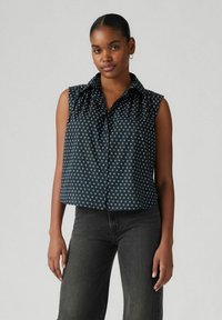 Blusa senza maniche blu navy con motivo floreale bianco, apertura frontale con bottoni e dettaglio del colletto arricciato. Abbinata a jeans scuri.