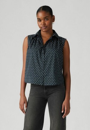 Blouse sans manches bleu marine avec motif floral blanc, devant à boutons et détail de col volants. Associée à un jean foncé.