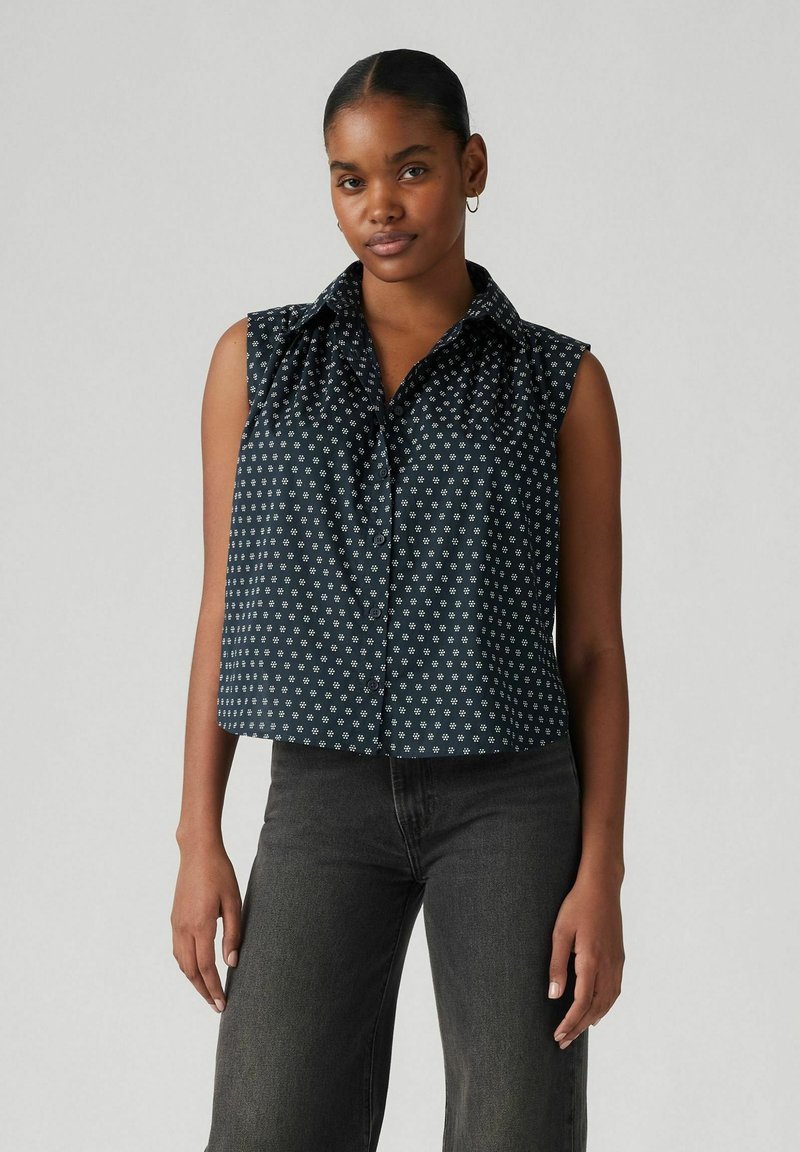 Blusa senza maniche blu navy con motivo floreale bianco, apertura frontale con bottoni e dettaglio del colletto arricciato. Abbinata a jeans scuri.