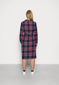 Abito a quadri blu navy con dettagli rossi e bianchi, di lunghezza media, vita con cintura, maniche lunghe e orlo dritto. Indossato con sneakers bianche.