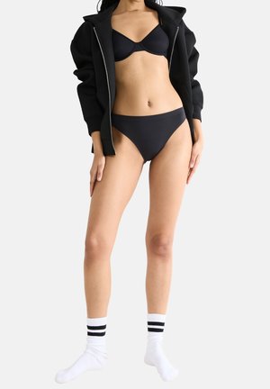 Ein schwarzer Sport-BH und passende Bikini-Unterwäsche werden unter einem schwarzen Zip-Hoodie getragen. Weiße Socken mit schwarzen Streifen runden das Outfit ab.