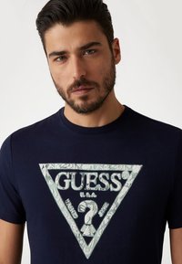 Navy t-shirt featuring a triangular green and white "GUESS" logo with textured patterns. Short sleeves, round neckline, cotton blend fabric.