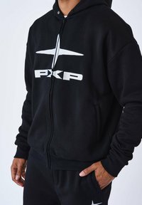 Schwarze Reißverschluss-Hoodie aus einem weichen Material, mit einem großen weißen "PKP"-Logo auf der Brust. Enthält eine Kapuze und seitliche Taschen.