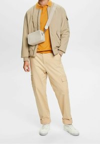 Beige Bomberjacke über einem orangefarbenen Polo, kombiniert mit cremefarbenen Cargo-Hosen und weißen Sneakers. Eine hellgraue Umhängetasche hängt von der Schulter.